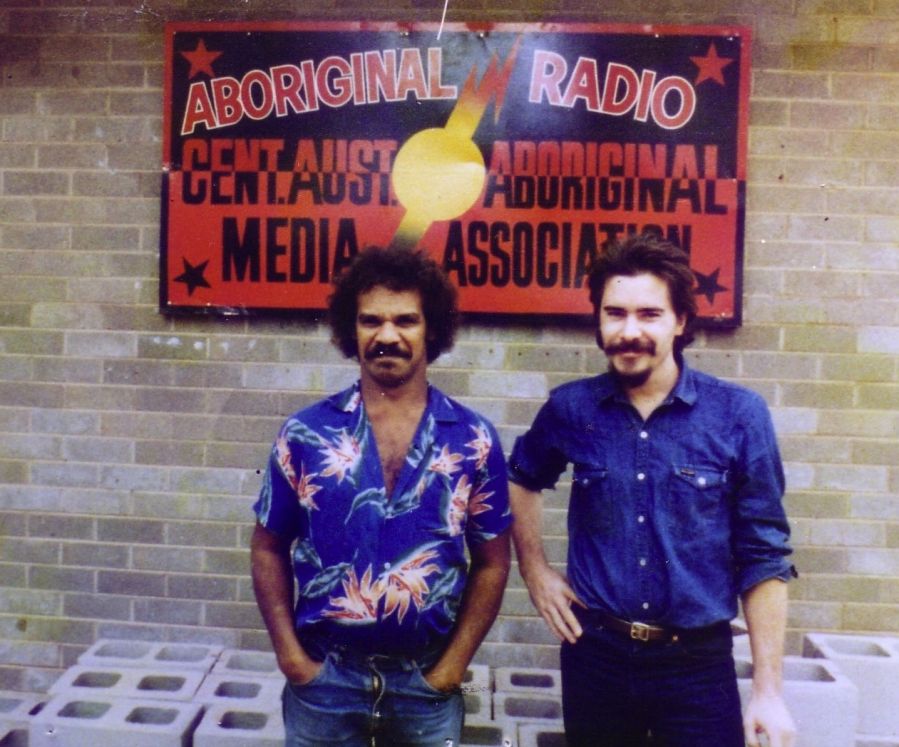 Photograph of two men in blue shirts standing in front of a sign reading Aboriginal Radio..