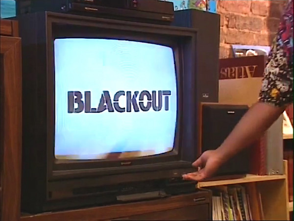 Screengrab. TV Screen. Blue background. Black title reading BLACKOUT. Woman reaches towards the TV buttons..
