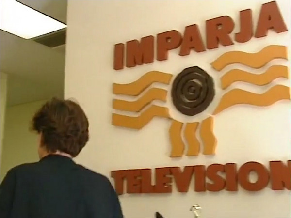 Screengrab. TV station. Logo on a wall, reading IMAPRJA TELEVISION in organe and yellow. Woman with brown hair walks past.