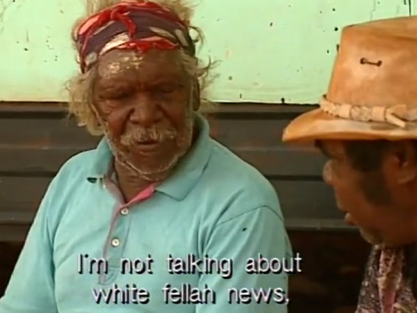Screengrab. Indigenous old man with beard in blue shirt is speaking to man in yellow hat. The subtitle reads - I'm not talking about white fellah news.