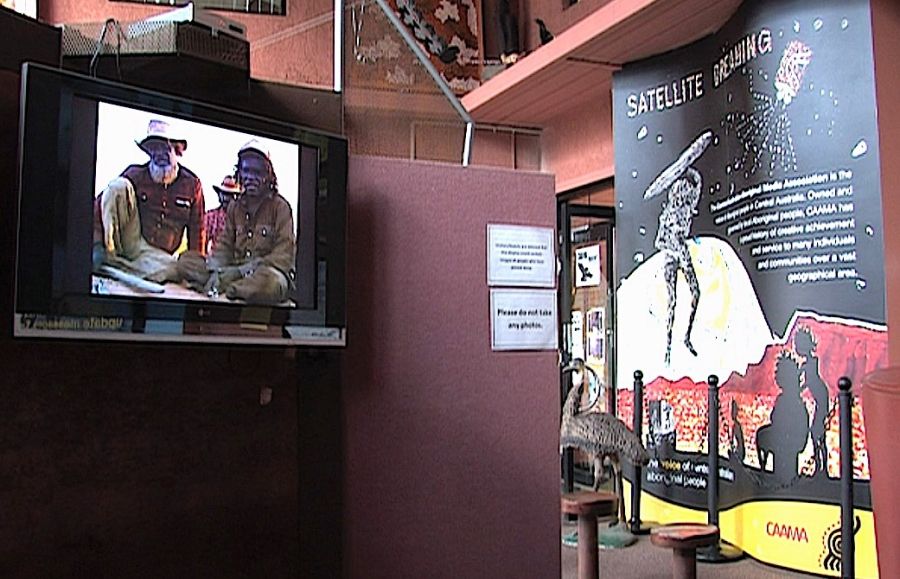 photograph. Interior. Television screen shows two men talking, a poster displays SATELLITE DREAMING in the background..