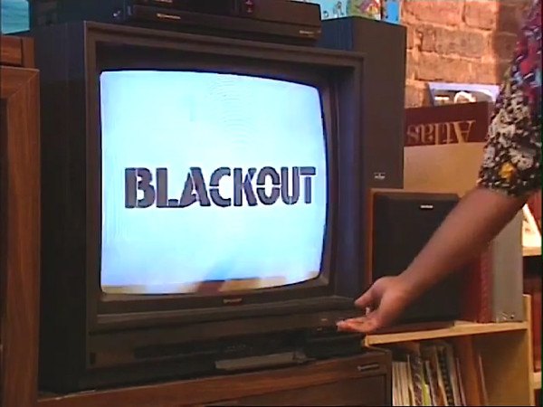 Screengrab. TV Screen. Blue background. Black title reading BLACKOUT. Woman reaches towards the TV buttons.